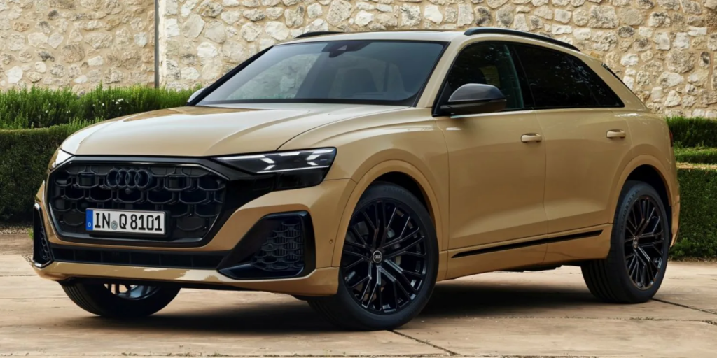 Ein goldfarbener SUV (Audi Q8) steht schräg von vorne fotografiert auf einer gepflasterten Fläche. Das Fahrzeug hat eine markante schwarze Front mit großem Kühlergrill, schmale LED-Scheinwerfer und schwarze Felgen. Im Hintergrund befindet sich eine helle Steinmauer mit etwas Grünbewuchs, was dem Bild eine ruhige, elegante Atmosphäre verleiht.