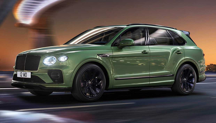 Ein grüner Bentley Bentayga SUV fährt auf einer Straße bei Abenddämmerung. Das luxuriöse Fahrzeug mit großen schwarzen Felgen und markantem Kühlergrill ist in Bewegung, während im Hintergrund eine beleuchtete Brücke verschwommen zu sehen ist.