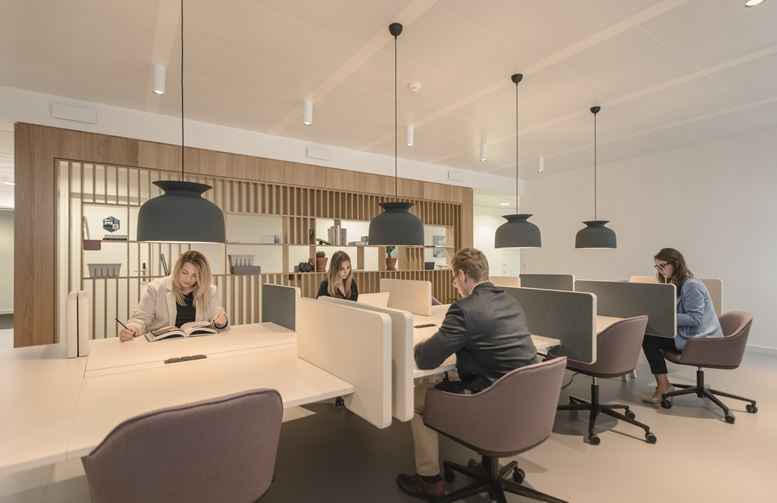 Ein moderner, heller Coworking-Space mit mehreren Arbeitsplätzen. Vier Personen sitzen an Tischen mit Trennwänden und arbeiten oder lesen, während große schwarze Pendelleuchten von der Decke hängen und eine Holzregalwand den Hintergrund bildet.