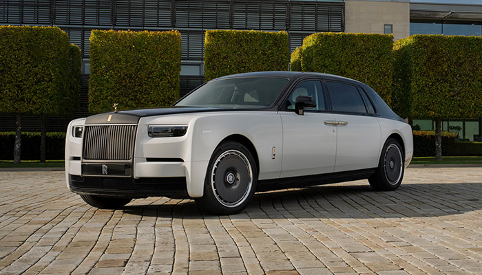 Eine weiße Rolls-Royce Phantom Limousine steht auf einem gepflasterten Vorplatz. Im Hintergrund befinden sich modern geschnittene Hecken und ein Gebäude mit großen Fenstern. Die Front mit dem markanten Kühlergrill und der Spirit-of-Ecstasy-Figur ist gut sichtbar.